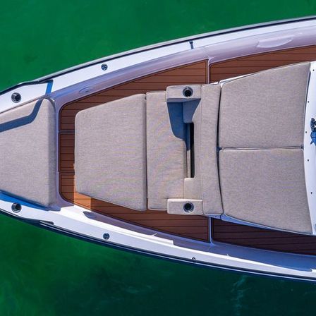 Aerial view of a white motorboat bow with teak decking and beige lounge cushions floating on emerald green water.