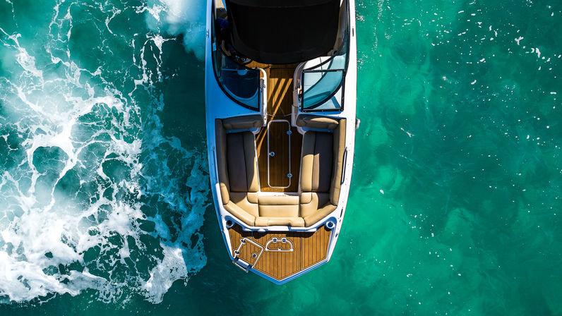 Aerial view of a white speedboat cruising turquoise waters, teak deck and cushioned bow seating with foamy wake and sunlit ocean