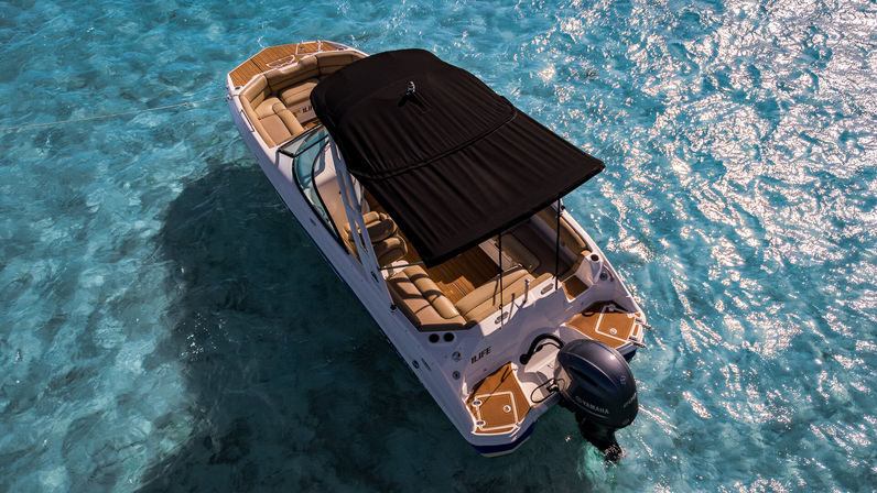 Aerial shot of a white motorboat with a black canopy, tan seating and outboard engine anchored in clear turquoise water with sunlight sparkling on gentle waves