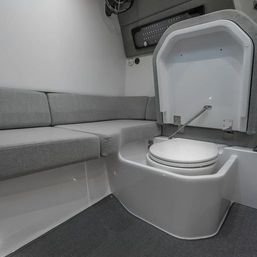 Compact boat cabin head with closed white marine toilet, lift-up access panel and gray L-shaped cushioned seating.