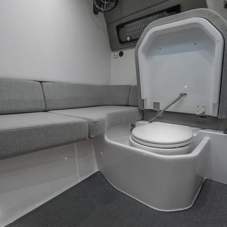 Compact boat cabin head with closed white marine toilet, lift-up access panel and gray L-shaped cushioned seating.