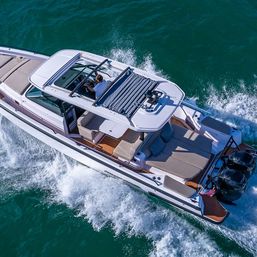Aerial view of a luxury white motorboat cruising turquoise coastal waters, open cockpit with driver, teak deck and aft sunpad lounge, twin outboard engines kicking up a foamy wake.