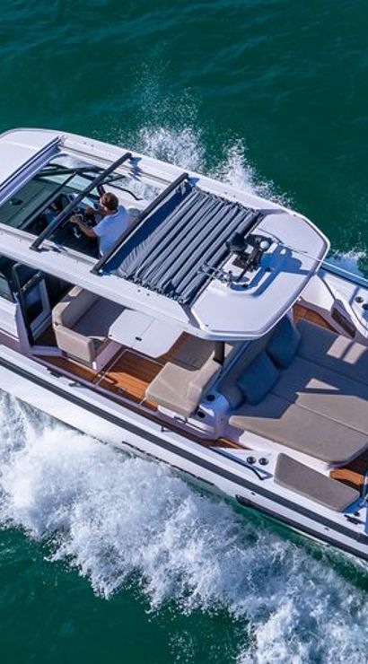 Aerial view of a luxury white motorboat cruising turquoise coastal waters, open cockpit with driver, teak deck and aft sunpad lounge, twin outboard engines kicking up a foamy wake.
