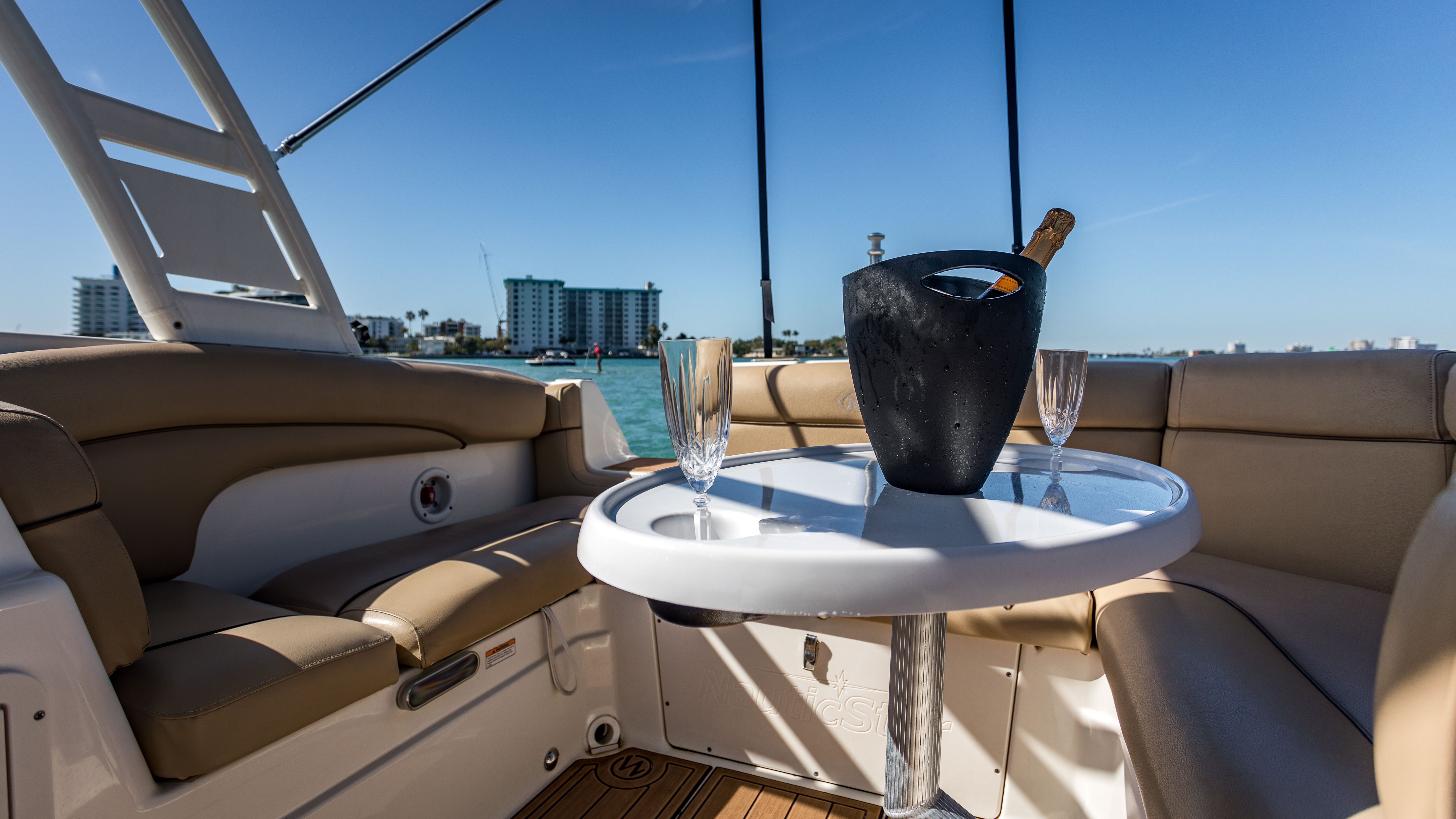 Sunlit yacht cockpit with tan leather seating and a white table holding a chilled champagne bottle in a black ice bucket and two flute glasses, turquoise bay and coastal buildings in the background, ready for a toast.