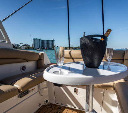 Sunlit yacht cockpit with tan leather seating and a white table holding a chilled champagne bottle in a black ice bucket and two flute glasses, turquoise bay and coastal buildings in the background, ready for a toast.
