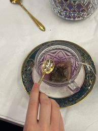 Close-up of a hand holding a gold teaspoon over a purple glass teacup with a steeping tea bag on an ornate blue-and-gold saucer on a white tablecloth