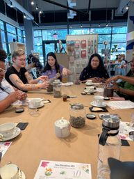 Tea blending workshop in a bright urban storefront: participants seated around a table with jars of loose herbs, teacups, spoons, a small honey jar and a printed 'Tea Blending 101' worksheet.