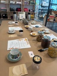 Tea tasting setup in a boutique: long table with vintage teacups, labeled jars of loose tea, menus, thermoses and snacks, folded apparel shelves in background
