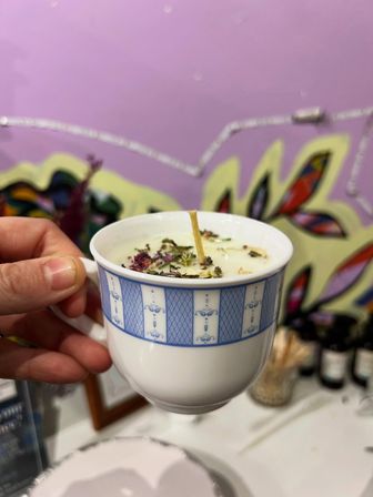 Hand holding a blue-patterned teacup candle with a wooden wick and dried flower petals, set against a bright purple mural background — cozy handmade soy candle close-up.