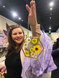 Smiling shopper lifts a hexagonal brass-framed glass suncatcher filled with pressed yellow and white daisies on a gold chain inside a colorful craft boutique.