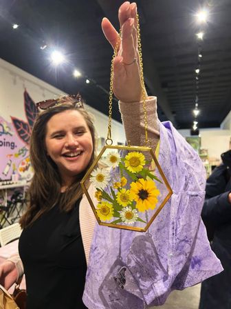 Smiling shopper lifts a hexagonal brass-framed glass suncatcher filled with pressed yellow and white daisies on a gold chain inside a colorful craft boutique.