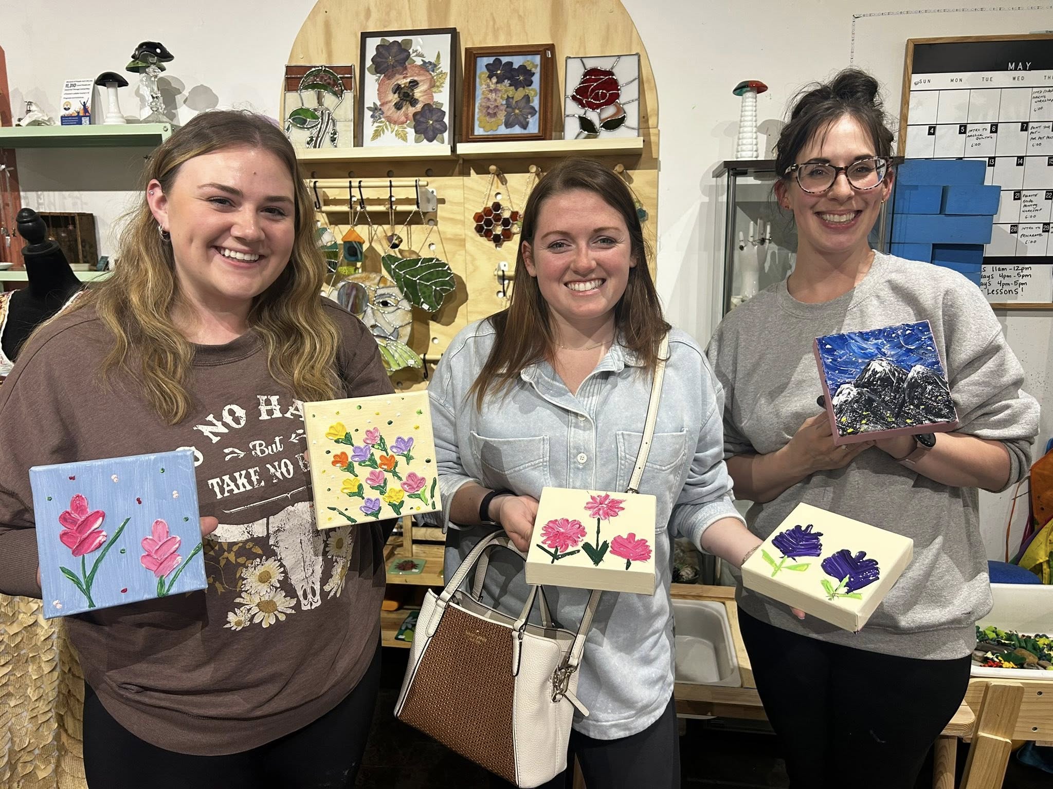 Three smiling women holding small painted canvases with colorful flowers and a mountain scene at a lively paint night in a cozy art studio, stained-glass artwork and supplies visible on shelves behind them.