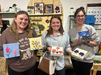 Three smiling women holding small painted canvases with colorful flowers and a mountain scene at a lively paint night in a cozy art studio, stained-glass artwork and supplies visible on shelves behind them.