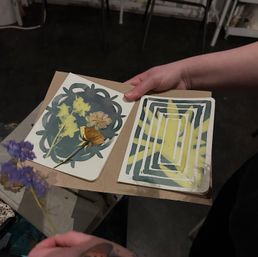 Hands holding an open kraft paper journal showing a pressed-flower botanical collage on one page and a layered yellow-and-teal geometric stencil print on the other, with a blurred purple flower in the foreground — DIY craft art journal.