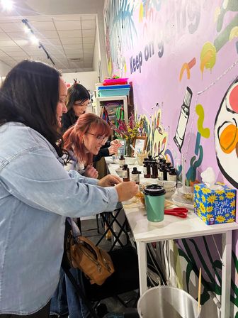 Three women at an indoor art studio workshop crafting at a table with small amber bottles, tools, a takeaway coffee cup and a bright colorful mural wall