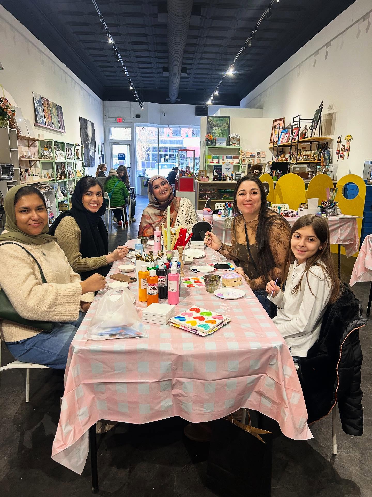 Five women and a girl smiling at a table in a bright community art studio, painting colorful crafts with brushes and acrylic bottles on a pink checkered tablecloth, shelves and storefront window visible in the background.