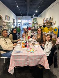 Five women and a girl smiling at a table in a bright community art studio, painting colorful crafts with brushes and acrylic bottles on a pink checkered tablecloth, shelves and storefront window visible in the background.