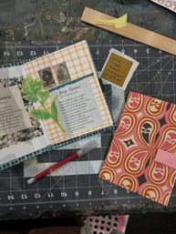 Top-down view of a craft workspace on a cutting mat with an open scrapbook spread featuring gingham paper, a botanical collage and poem, a patterned paper envelope, and a red craft knife.