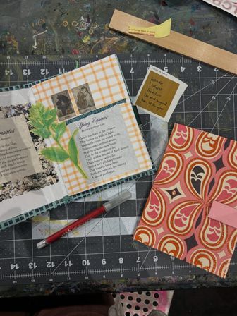Top-down view of a craft workspace on a cutting mat with an open scrapbook spread featuring gingham paper, a botanical collage and poem, a patterned paper envelope, and a red craft knife.