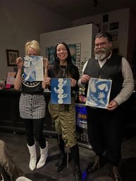 Three people in an indoor art studio proudly holding blue-and-white cyanotype botanical prints after a printmaking workshop