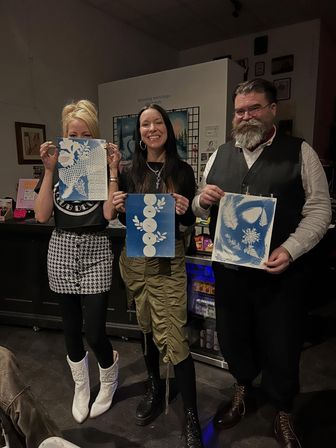 Three people in an indoor art studio proudly holding blue-and-white cyanotype botanical prints after a printmaking workshop