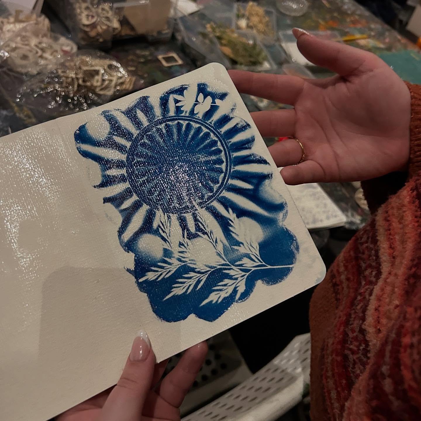 Close-up of hands holding a white canvas panel featuring a blue stenciled sunburst and leafy botanical design on a craft table with supplies — DIY stencil art project.