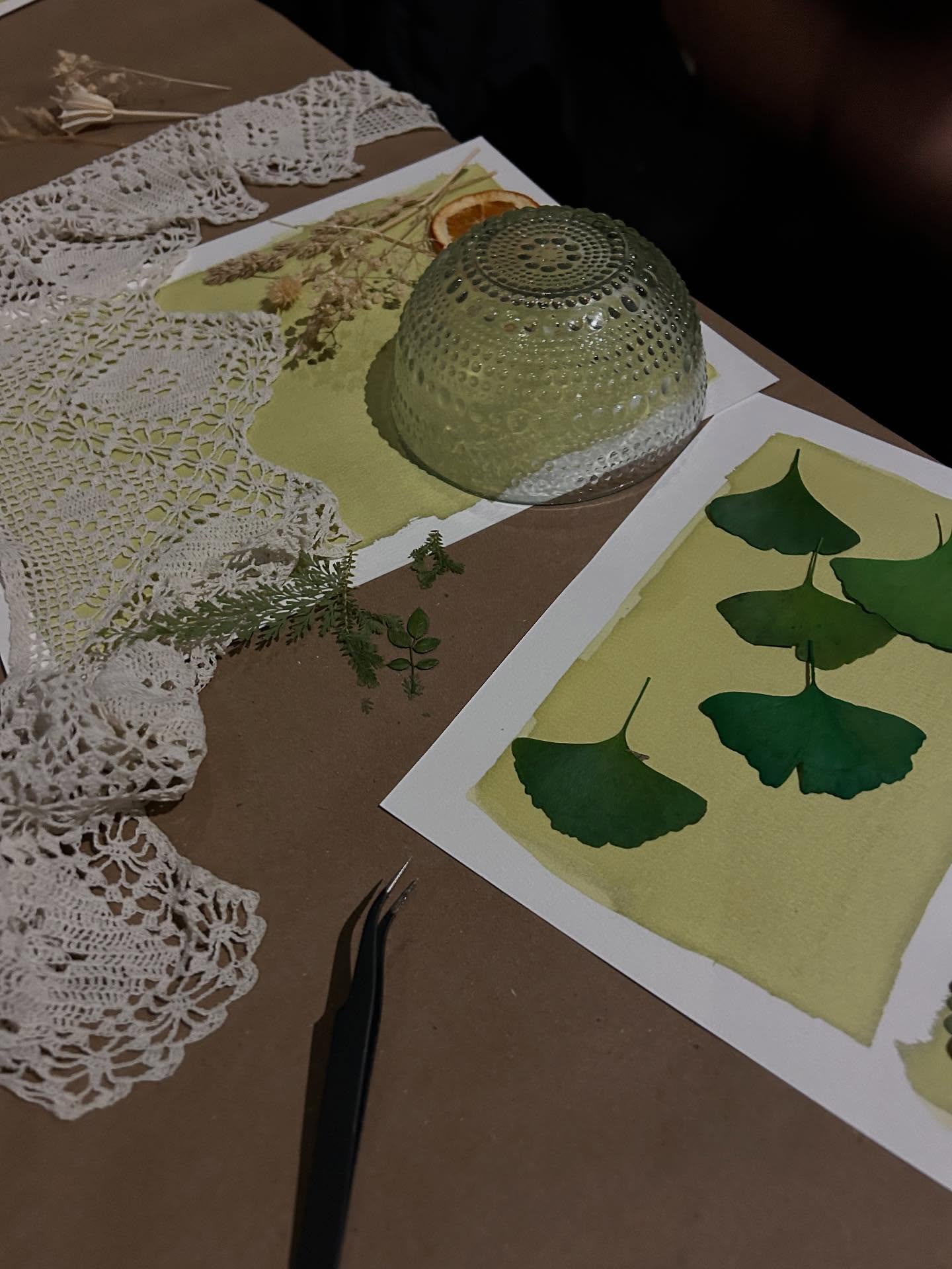 Craft table with ginkgo leaf botanical prints on watercolor paper, crochet doily, tweezers, dried flowers and a textured glass bowl — DIY nature art setup.