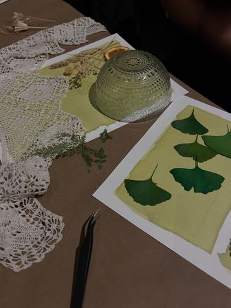 Craft table with ginkgo leaf botanical prints on watercolor paper, crochet doily, tweezers, dried flowers and a textured glass bowl — DIY nature art setup.