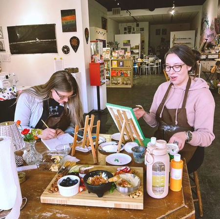 Two people at a community art studio painting on tabletop easels during a paint night, wooden table with canvases, paints, brushes and a charcuterie-style snack board, craft shelves and seating in the background.