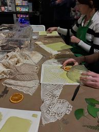Indoor DIY craft workshop table with vintage lace doilies, dried orange slices and green paper, hands using a glass bowl for eco-printing, assorted lace trims and craft supplies