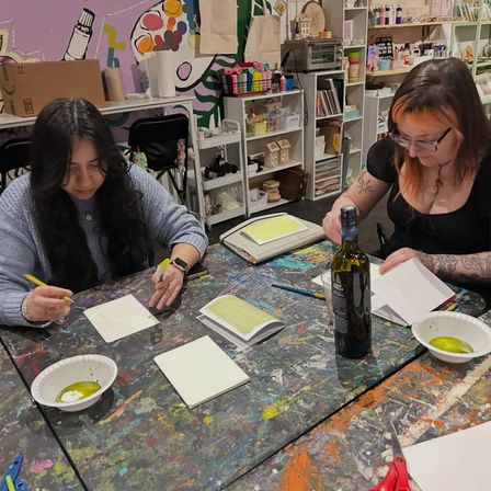 Two people painting at a paint-splattered communal table in a colorful indoor art studio, working on watercolor papers with brushes, bowls of green paint and a wine bottle nearby.