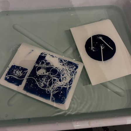 Two DIY cyanotype sunprints floating in a shallow tray: a rectangular print with tangled white string patterns over deep blue and a circular blue photogram with three white lines, on small white paper sheets.