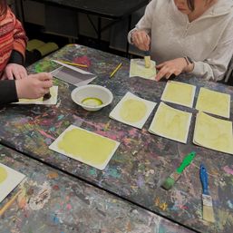 Hands painting yellow watercolor shapes on small cards at a paint-splattered art studio table with brushes, pencils and a bowl of paint — casual craft workshop scene