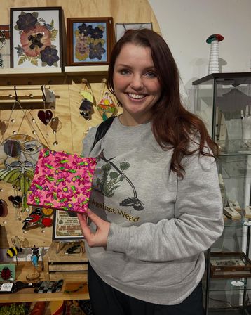 Smiling shopper in a gray sweatshirt holding a bright pink hand-painted mosaic tile inside a local artisan gift shop with stained-glass art, jewelry and glass display cases