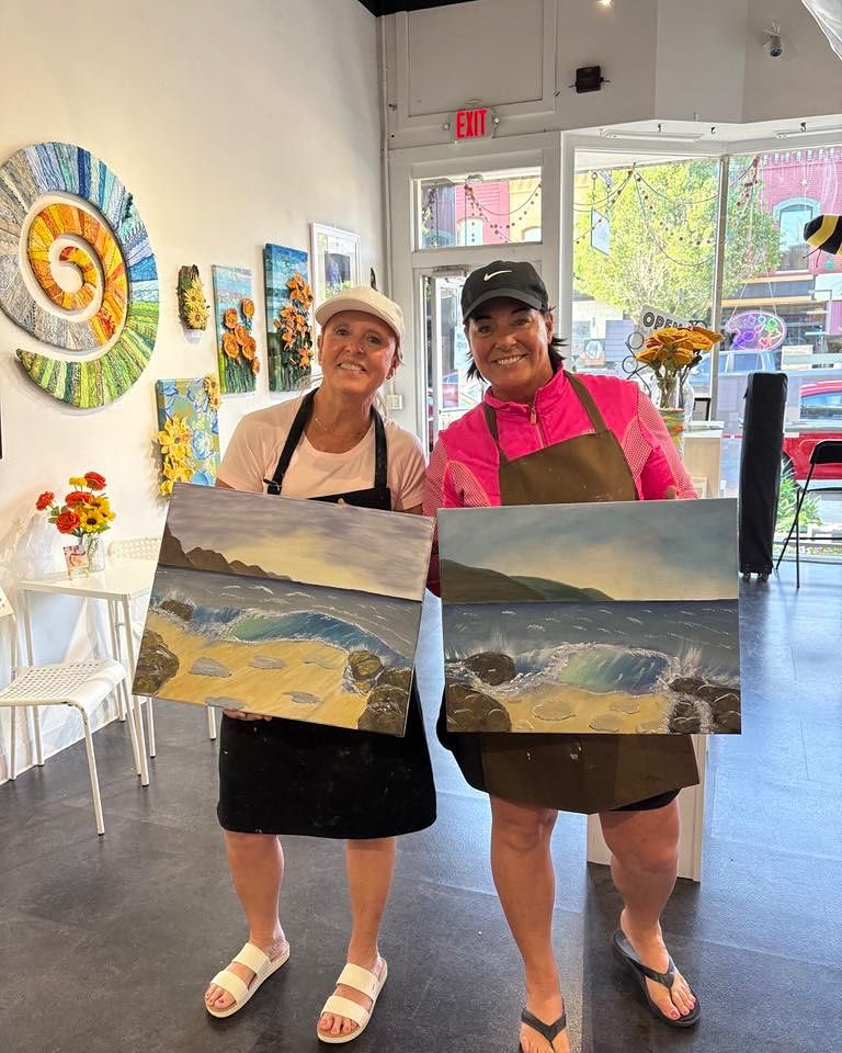 Two women in aprons holding matching seascape paintings in a bright downtown art studio, colorful wall art and sunflowers on display with street-facing windows in the background.