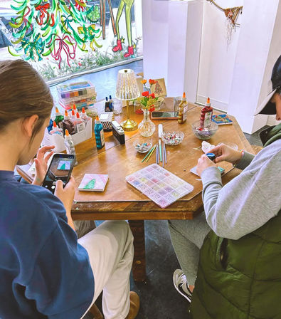 Cozy holiday craft workshop at a wooden table — two people making small Christmas canvases with glue bottles, glitter, beads, paintbrushes and a bead organizer, beside a storefront window painted with festive ribbons and trees.