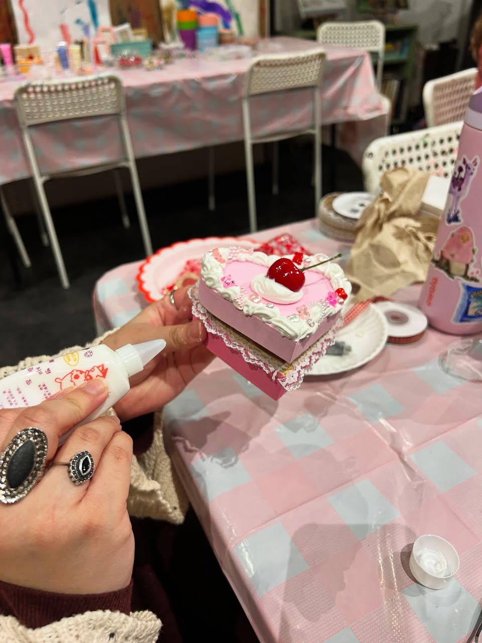 Hands applying glue to a pink heart-shaped DIY box decorated with faux whipped cream, a cherry, beads and lace on a pink-checkered craft table at a craft workshop.