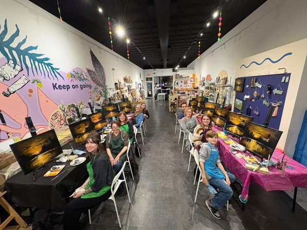 Wide-angle shot of a lively indoor painting workshop in an art studio — rows of easels with sunset canvases, participants at long tables, colorful mural reading “Keep on going” on the wall.
