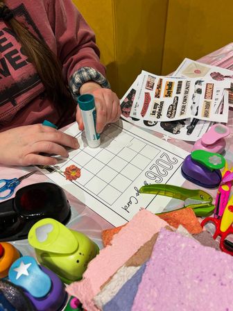 Person's hands gluing a flower sticker onto a 2026 DIY calendar grid at a craft table surrounded by colorful paper punches, scissors, sticker sheets and textured handmade paper.