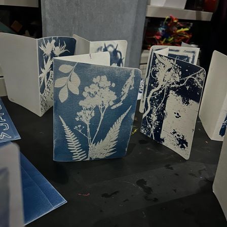 Mini cyanotype notebooks with white botanical silhouettes and abstract prints on blue covers, displayed on a black craft table in an art studio.