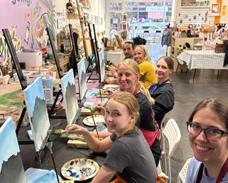 Smiling group painting at a bright community art studio, seated at a row of easels with mountain-and-sky canvases, palettes, brushes, and a storefront window in the background.