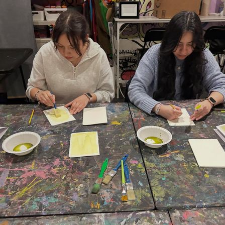 Two people at a paint-splattered art studio table working on small watercolor cards, with bowls of green paint, sponges and brushes scattered across the workspace.