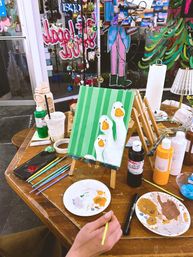 Tabletop painting scene at a community art studio: small easel with a canvas of three white geese on green stripes, surrounded by paint bottles, brushes, palettes, and a hand holding a thin brush with a colorful window mural in the background.