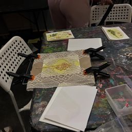 Paint-splattered art studio table with a DIY greeting card clamped flat — lace trim and dried pressed flowers on yellow paper, black spring clamps, stacks of blank white cards and botanical cards, white perforated chairs nearby.