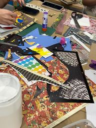 Overhead view of a busy craft table in a studio with patterned scrapbook papers, colorful stickers, scissors, glue stick and hands assembling a collage