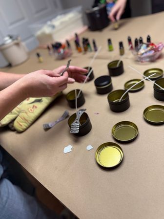 Hands placing wicks into matte black metal candle tins on a brown paper–covered table during a DIY candle-making workshop, with wick clips, small essential oil bottles and an oven mitt visible.