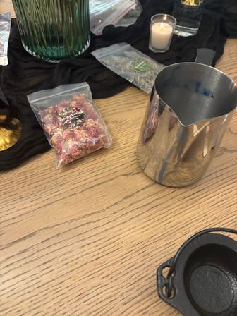 Light wood tabletop display with a reflective stainless milk pitcher, small cast-iron pot, resealable bag of dried pink flowers, lit votive candle and green glass vase on black cloth.