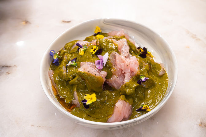 Gourmet seafood bowl of thinly sliced raw white fish draped in vibrant green herb sauce and dotted with colorful edible flowers, served in a white bowl on a marble countertop.