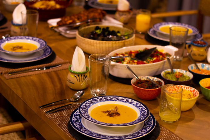 Cozy wooden dining table set for a family-style meal with blue-patterned bowls of creamy orange soup, colorful bowls of salsas and dips, a salad, drinking glasses and napkins.