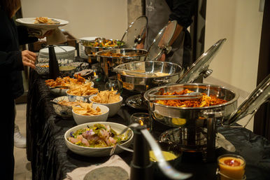Indoor catered buffet at a reception with stainless-steel chafing dishes of hot entrees, bowls of tortilla chips, fresh salads and sides arranged on a black tablecloth as guests serve themselves.
